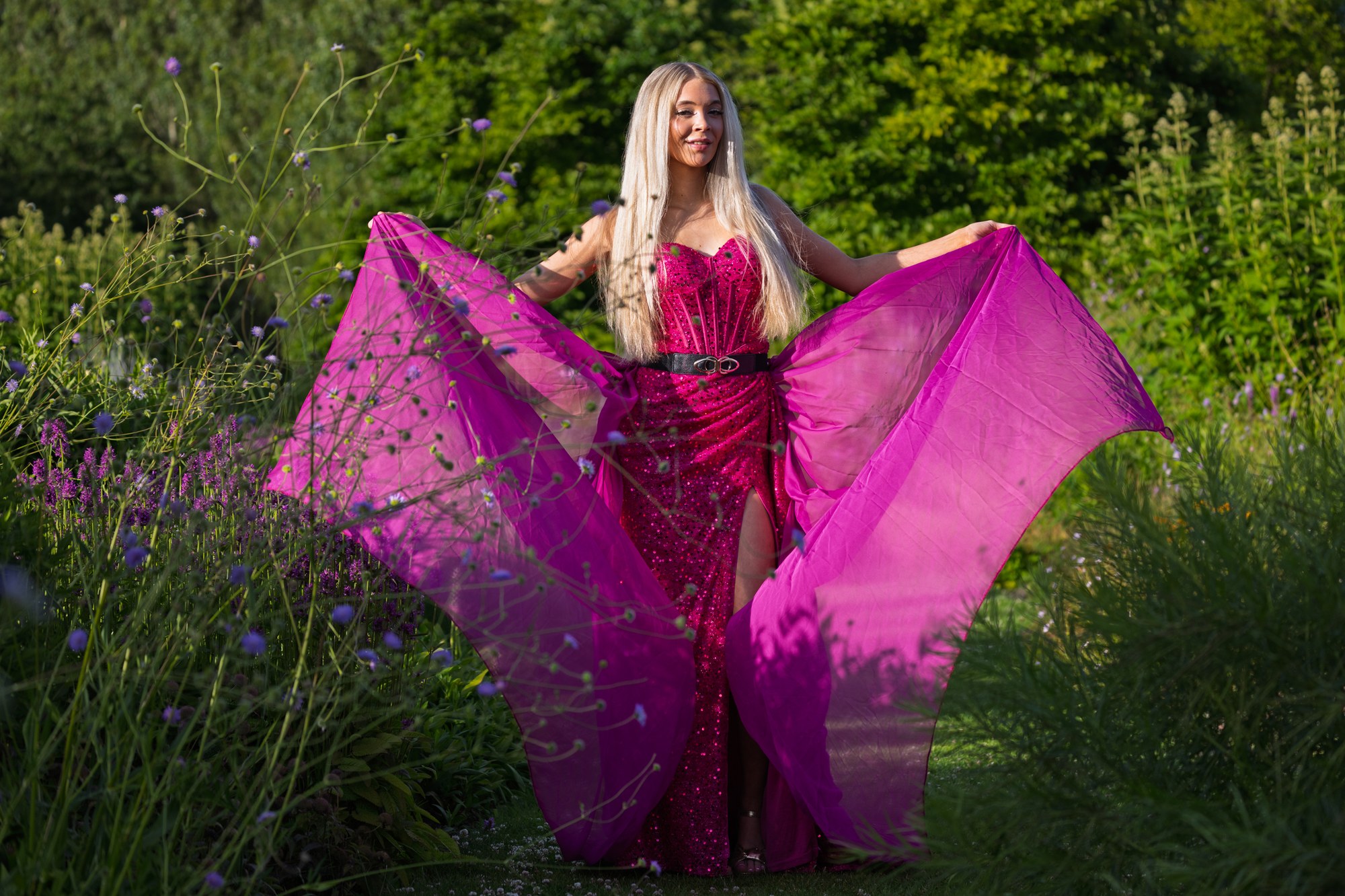 Model spreidt de paarse sleep van haar fuchsia jurk als vleugels tussen wilde bloemen in laag avondlicht, Maximapark Utrecht