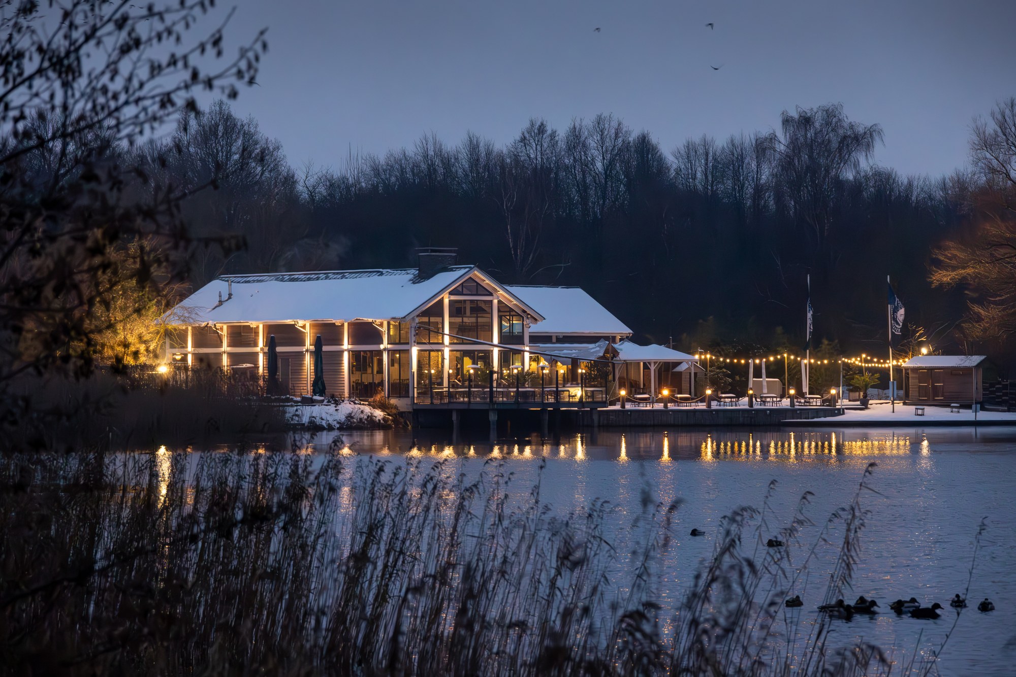 Verlicht paviljoen aan een besneeuwd meer in het blauwe uur, warme lichtjes weerspiegeld in het water, Nederland