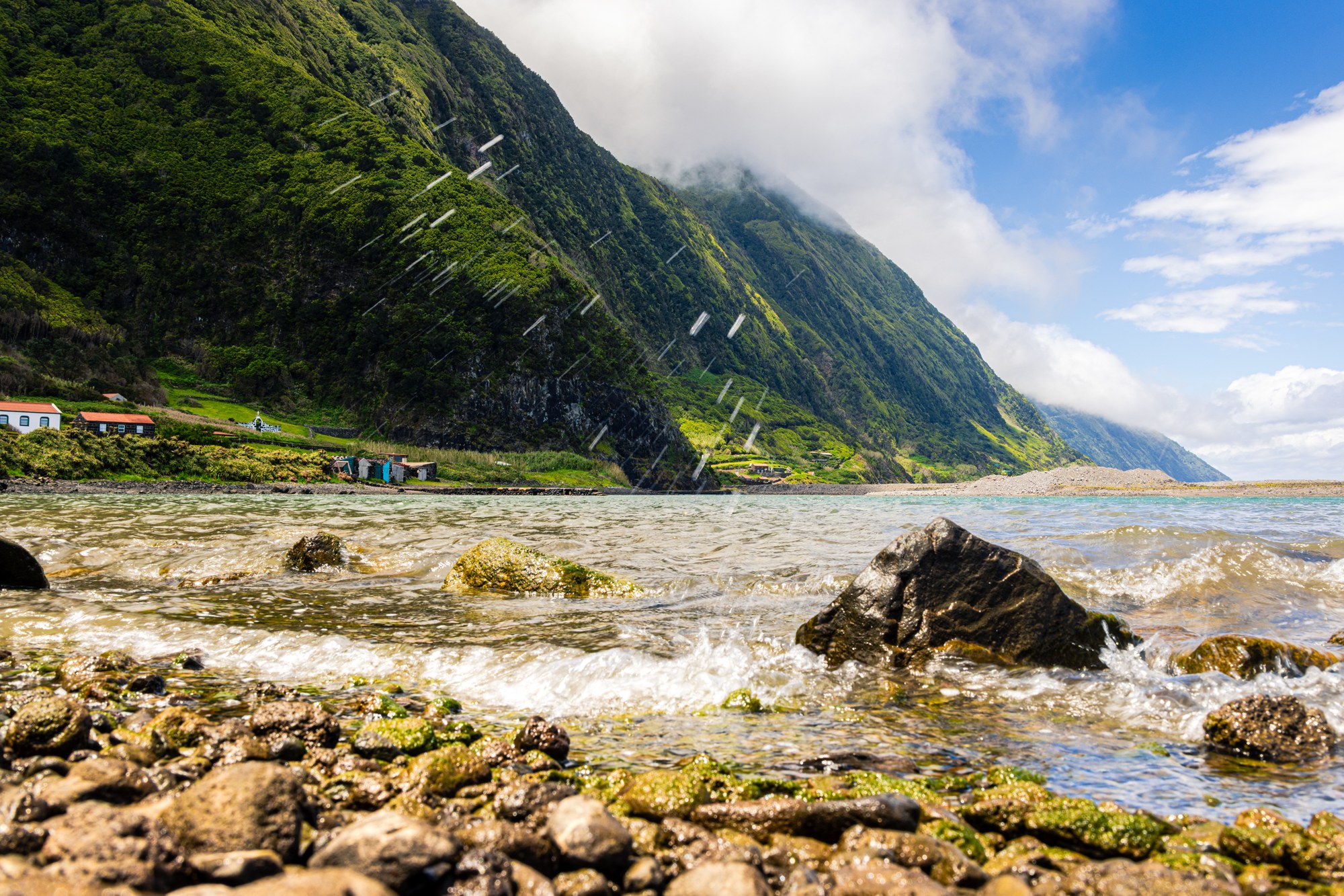 Brekende golven over bemoste rotsen op de fajã-kust, oceaanspray in de lucht, groene klifwand en turquoise baai, Azoren