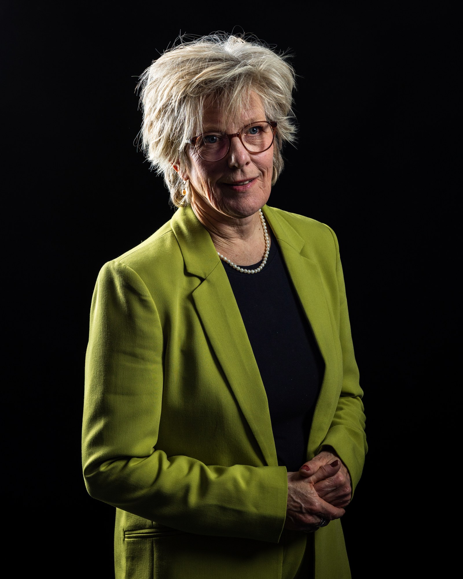 Oudere vrouw in geelgroene blazer met parelketting, milde glimlach, studioportret op zwart