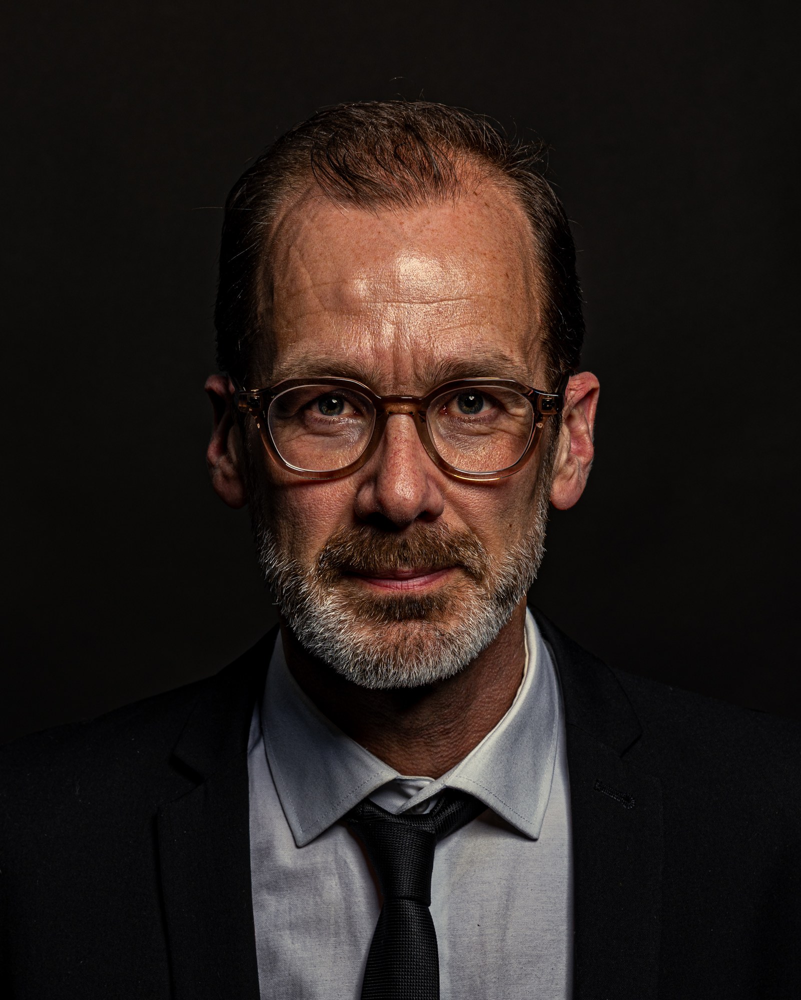 Man met transparant montuur die recht in de lens kijkt, studioportret op zwart
